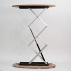 A wooden table with a round top is supported by a silver, scissor-lift mechanism for adjustable height. The base and tabletop have a natural wood finish, set against a plain white background.