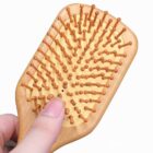 A person holding a wooden paddle hairbrush with round-tipped bristles. The brush features a light wood finish and closely spaced pins. The background is plain white.
