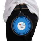 Person wearing black pants with a blue collapsible bowl attached to their belt by a carabiner. The bowl has a label with a heart logo and the word Volunteer.