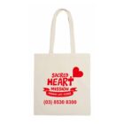 Canvas tote bag with red text and graphics. Reads: Sacred Heart Mission with a heart and banner stating Making Life Easier. Includes a phone number: (03) 8530 8300. Bags handles are visible above.
