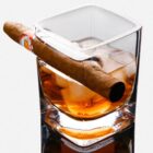 A glass of amber-colored liquor with ice cubes, topped with a brown cigar resting on the rim, against a plain background.