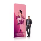A man in a suit stands next to a large pink sign featuring a woman in athletic wear jumping. The sign has the text LETS GO! and additional text at the bottom. The sign contrasts with the mans formal appearance.