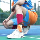 Person crouching on a sports court, wearing a colorful outfit with a blue and orange jersey, shorts, and bright patterned socks. They have white sneakers and are in an outdoor setting with a green fence behind them.