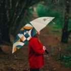 A person in a red coat stands on a forest path, holding an umbrella with a colorful geometric pattern. The path is surrounded by tall trees and the atmosphere appears peaceful and slightly overcast.