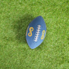 A blue American football with white stitching and yellow logos, including the text Infinity Design, rests on a vibrant green grass field.