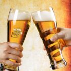 Two hands each hold a tall, curved beer glass, clinking them together. One glass is labeled Beer and the other Craft Beer. The background is a warm, golden gradient, enhancing the festive and celebratory feel.