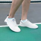 A person wearing white sneakers with light blue socks is walking on a green and gray surface, possibly a sports court. The shoes have a sleek design with a black accent on the heel.