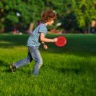 Child with curly hair wearing a striped shirt and jeans is playing with a red toy in a lush green park. Trees are visible in the background under a sunny sky.