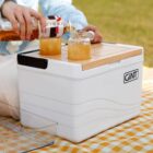 A person is pouring a drink from a clear bottle into two glass jars on a picnic tablecloth. The jars are placed on a white cooler with a wooden lid. The cooler has the brand name GINT on it.