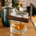 A glass of whiskey is placed on a wooden surface with a cigar resting on top. Theres a metal tumbler with blurred text in the background, and soft, warm lighting creating a cozy atmosphere.