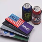 Two beverage cans are in foam koozies, one black with Woodford National and the other red with DET. A stack of more koozies lies nearby, featuring designs like an American flag, a soccer ball, and a casino logo.