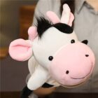 A person holds a soft, plush toy cow with pink ears and snout, black spots, and black hair tuft. The cow has a smiling face and is being used as a puppet against a blurred indoor background.
