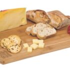 A wooden serving board with a block of yellow cheese, sliced bread, strawberries, crackers, and cheese cubes.