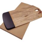 A wooden cutting board with a smooth, dark edge and a leather strap in a corner sits on top of a flat, brown cardboard box.