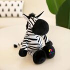 A plush zebra toy with black and white stripes, black hooves, and a fluffy mane sits on a cream-colored surface. The right hoof features a colorful Kidsplace logo. The background is blurred with a hint of green leaves and a patterned pillow.