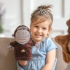 A young girl wearing a tiara and a blue dress smiles while holding a monkey hand puppet. She is seated on a couch, and the background is softly blurred.