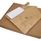 A bamboo cutting board shaped like a house with a white rectangular dish and a small cheese knife on top, placed on a plain brown cardboard box.