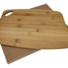A rectangular bamboo cutting board with a small handle cut-out rests on a plain brown cardboard box. The board has a light wood grain finish with subtle color variations.