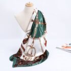 A silky scarf with a rich pattern drapes elegantly over a mannequin bust. The design features chains, straps, and ornate details in green, gold, and brown tones. A colorful magazine lies partially visible in the background on a white surface.