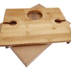 A wooden wine and cheese board with slots for glasses and a center hole, resting on a closed cardboard box. The board has a smooth, natural finish.