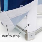 Close-up of a fabric corner secured by a Velcro strip, with an arrow pointing to the strip. The fabric has a blue and white pattern, and its attached to a metal frame. The image demonstrates how the Velcro is used for fastening.