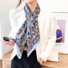 A person wearing a white blazer and a patterned blue scarf stands casually. Theyre holding sunglasses and have a small white bag nearby. A framed photograph of a person in a blue top and hat is in the background.