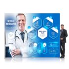 Smiling doctor in a white coat stands next to a medical infographic display. The infographic features blue hexagonal icons labeled Medication, Medical Box, Call 24/7, and Check Up. A man in a suit stands to the right of the display.