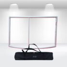 A white metal frame is set against a white background under two round spotlights. Below the frame is a black carrying bag with handles, placed on a light gray surface.