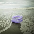 A purple inflatable chair floats on shallow water at the edge of a beach. The sky is overcast, and waves are gently rolling in.