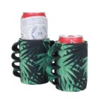 Two beverage cans are in green and black neoprene holders with finger grips, featuring a palm leaf design. The holders enable easy handling and provide insulation for the drinks.