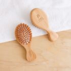 Two small wooden hairbrushes with brown bristles, one facing up and the other down, rest on a light wooden surface. They have engraved text and are placed on a white towel. The scene has a minimalist aesthetic.