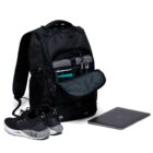 A black backpack with multiple compartments is open, revealing organized items like a phone, notebooks, and pens inside. Nearby are a pair of black and white sneakers and a closed laptop.