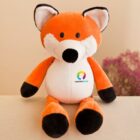 A plush toy fox with an orange body, white face, and black ears and paws sits on a beige surface. The fox has a colorful logo on its chest. In the background, there is a blurred pink and white floral arrangement.