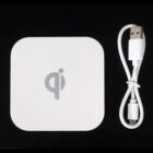 A white square Qi wireless charging pad with rounded corners is placed next to a coiled white USB cable against a black background.