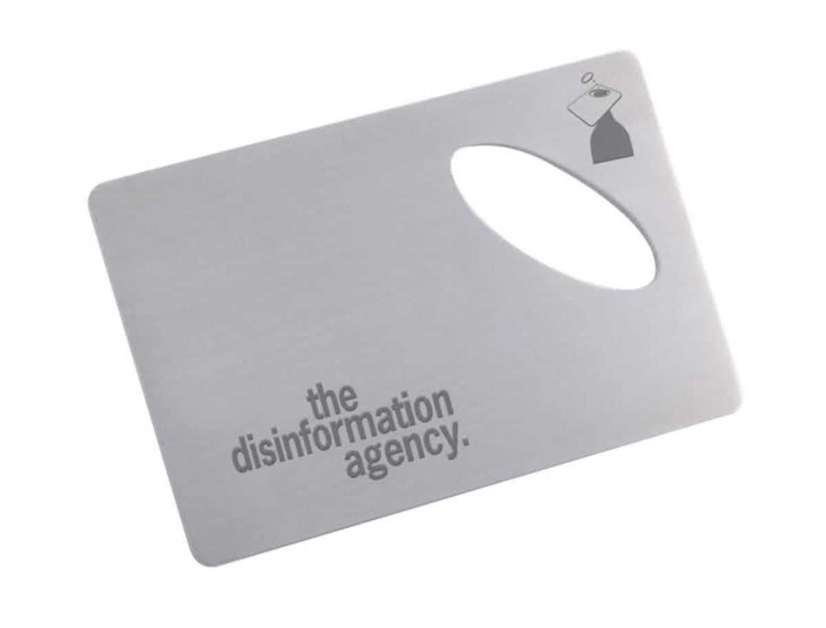 A metal card with an oval cutout features the text the disinformation agency in bold, black letters. A small illustration of a TV silhouette with a hat is in the top right corner.