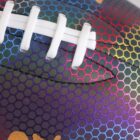 Close-up of an American football featuring a colorful hexagonal pattern with a rainbow gradient. The white laces contrast against the vibrant background.
