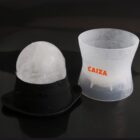 A transparent plastic ice mold with an orange CAIXA logo next to a similar black mold. The molds are used to create a perfectly round ice sphere, which is visible sitting in the black mold on a dark surface.