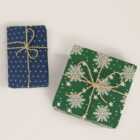Two gift boxes wrapped in festive paper. The smaller box has dark blue wrapping with small white polka dots, while the larger box features green paper with white snowflakes and stars. Both are tied with rustic twine bows.