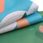 A close-up of folded fabric layers featuring abstract patterns in blue, green, peach, and white colors. The fabric texture is visible, and each layer is neatly stacked, showcasing the vibrant design elements.