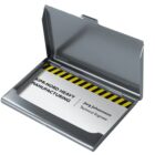 A silver business card holder is open, displaying a card with the text Kirr-Nord Heavy Manufacturing and Jorg Johansson, Technical Engineer alongside a yellow and black striped design.