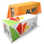 Two tables covered with custom tablecloths. The front tablecloth is white with colorful red, yellow, and green shapes and curacao text. The back tablecloth is orange with BMG Marketing Group text and a logo on one side.