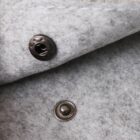 Close-up of a gray wool fabric with a metal snap button. The top piece with a hole is attached to the fabric above, while the bottom half is sewn below, creating a fastening mechanism. The fabric has a soft, felt-like texture.