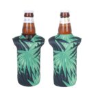 Two beer bottles are shown, each inside a neoprene sleeve with a tropical leaf pattern in shades of green. The sleeves have a V-shaped opening at the neck, exposing the bottle caps. The labels on the bottles are partially visible.