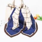 A white and navy blue scarf with brown edges draped over a white handbag. Nearby, two orange tulips and a green stem lie on a white surface, alongside a partially visible card with black text.