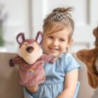 A young girl with a tiara smiles while holding a brown and beige plush puppet shaped like an animal. She is sitting on a couch, wearing a light blue dress. Another puppet is partially visible in the foreground.