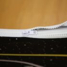Close-up of a white zipper partially opened against a black and yellow abstract background, possibly representing space with stars. The zipper is at the top of the image, emphasizing its teeth and slider.