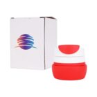 A red and white collapsible cup with a lid is displayed next to its packaging. The white box features a colorful logo on the front. The cup is compact, emphasizing its portable design.