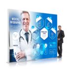 A doctor in a white coat stands smiling with arms crossed next to a large medical infographic display. The infographic features icons for medical services such as check-ups and medication, along with hexagonal designs and text.