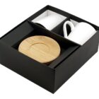 A black gift box contains two white mugs and two wooden coasters arranged neatly inside. The wooden coasters are stacked together, and the mugs are placed side by side in separate compartments.