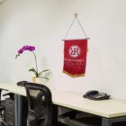 An office setting with two desks, each with a black chair and a phone. A potted orchid plant is placed on the left desk. A red wall hanging with the text Witherspoons River Run is displayed on the light-colored wall.